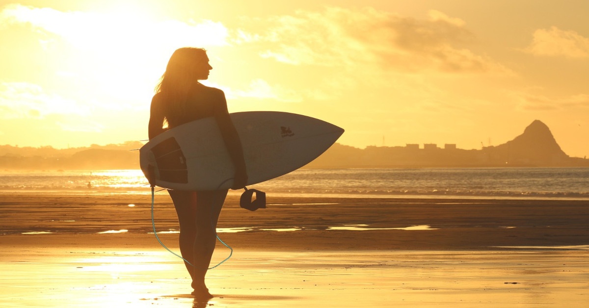 A surfer is standing in the sunlight on the beach with a surfboard.