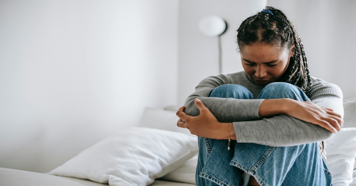 A woman who is suffering from depression is sitting on her bed while looking sad and holding her legs.