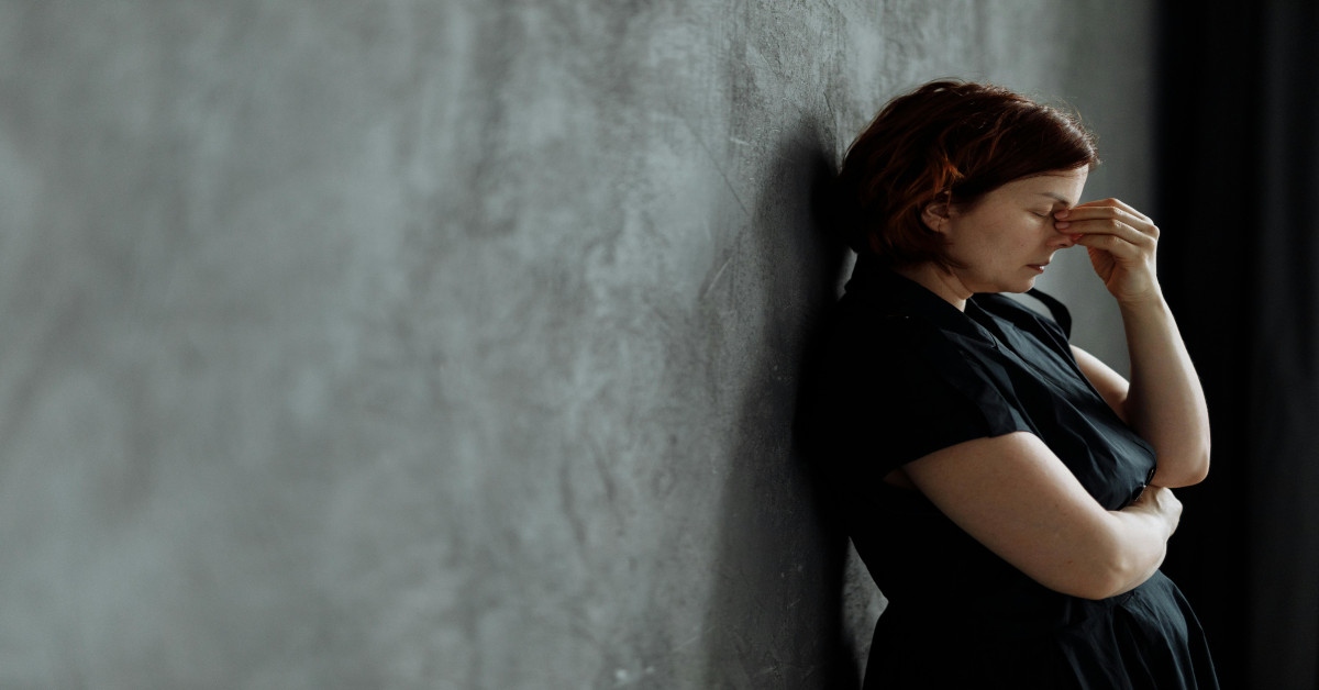 A woman leaning against a wall with her back while holding her head and looking sad.