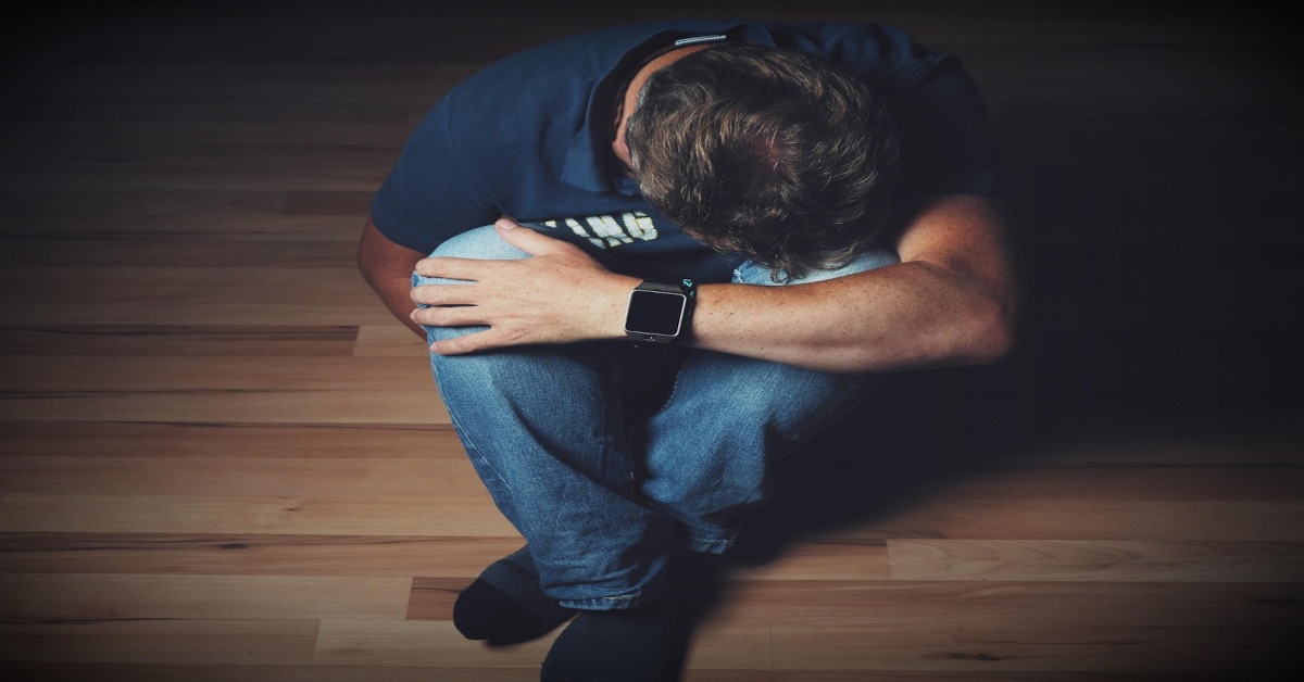 A depressed man sitting on the floor while holding his legs with his head down.
