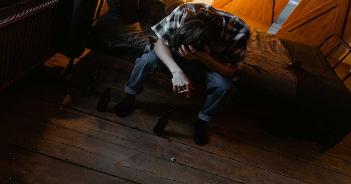 A distressed man is smoking a cigarette while sitting on his bed and holding his head.