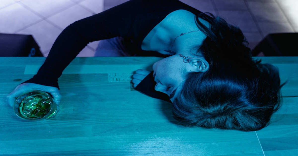A drunk woman holding her drink while lying passed out drunk on the bar.