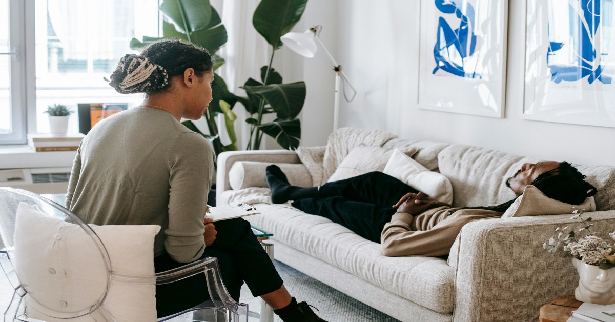An ethnic man is lying on his back on a couch while speaking to a psychologist.
