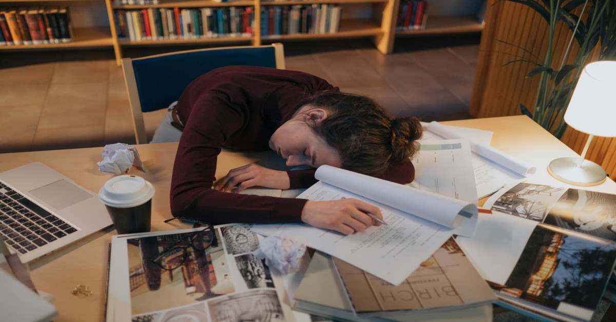 An exhausted woman sleeping on her desk between books.
