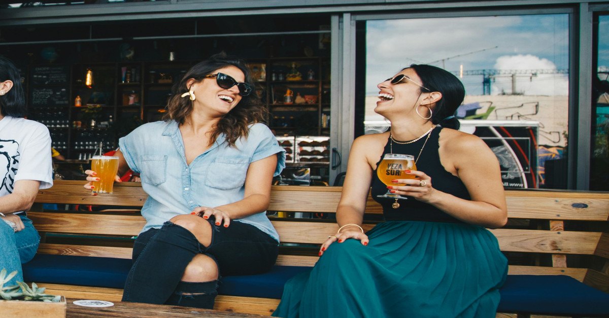 A group of females is laughing and drinking together on a bench outside.