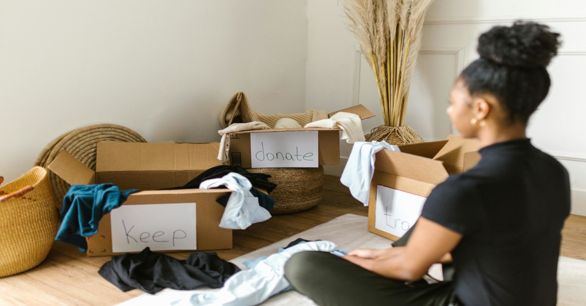 A person sitting in front of three boxes with clothes reading "keep", "donate", and "trash".