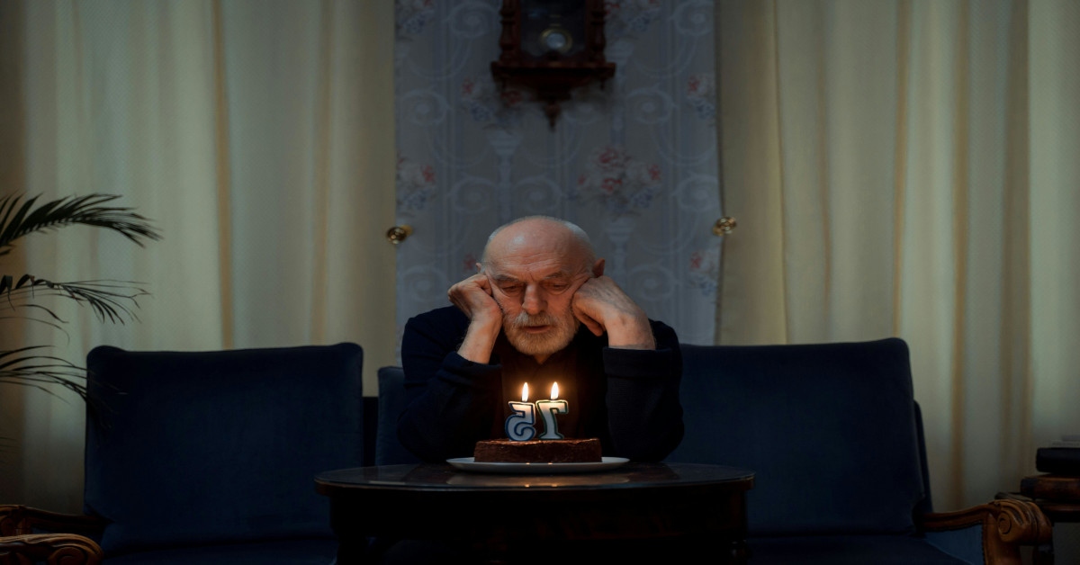 A lonely man sitting alone on his couch while blowing out the candles of his birthday cake for his 75th birthday.