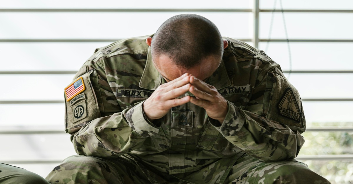 A man in the army looking down while holding his head.