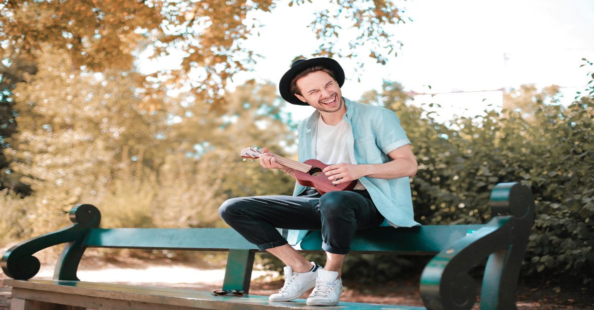 Image of a man playing an instrument on a bench outside while laughing.