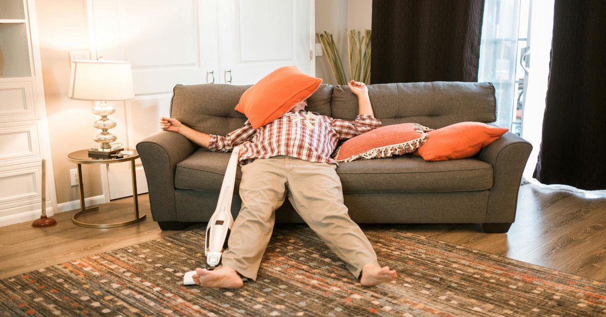 Man is lying on the sofa with his face covered with a pillow with a vacuum beside him.