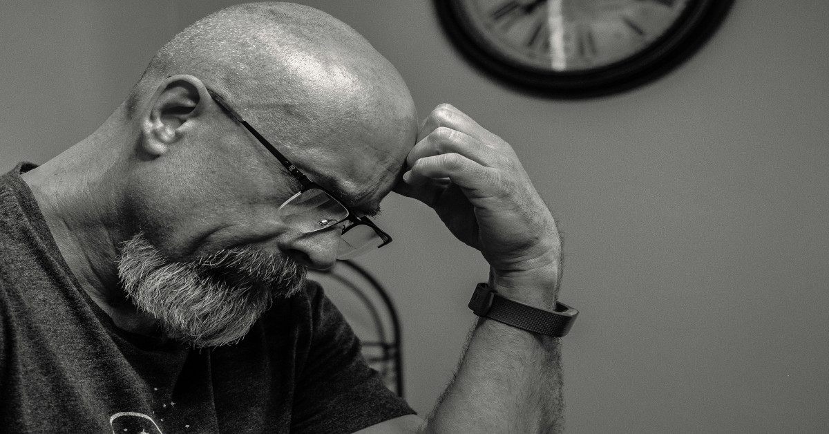 A black and white image of a man thinking while holding his head in front of a clock.