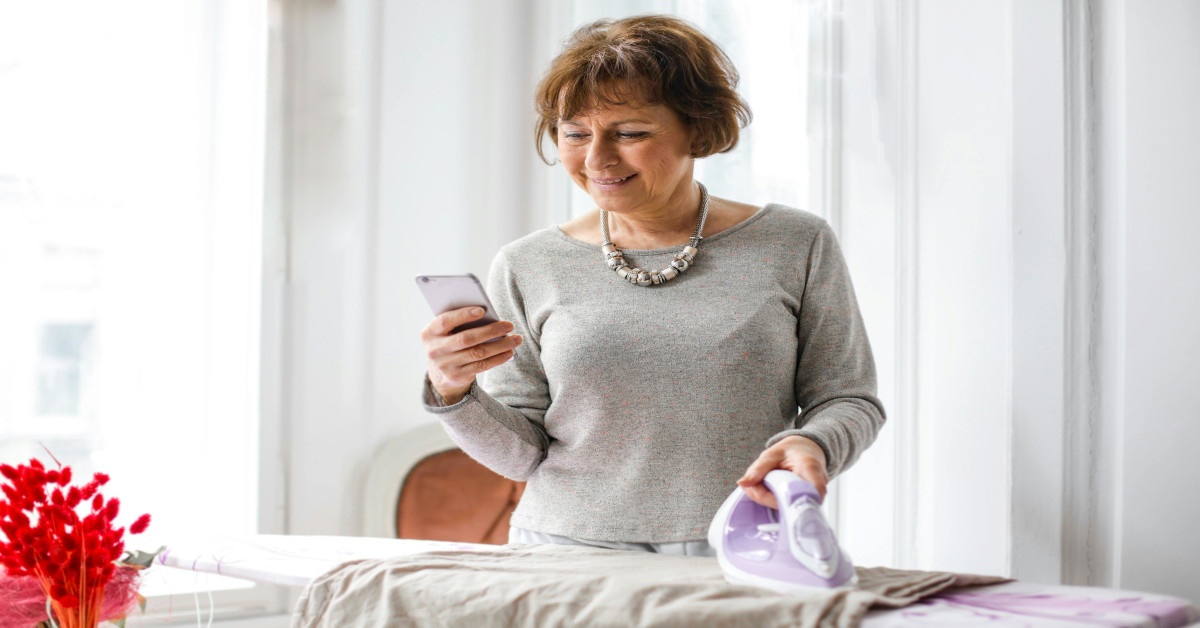Image of a middle-aged woman smiling while ironing and looking at her cellphone.