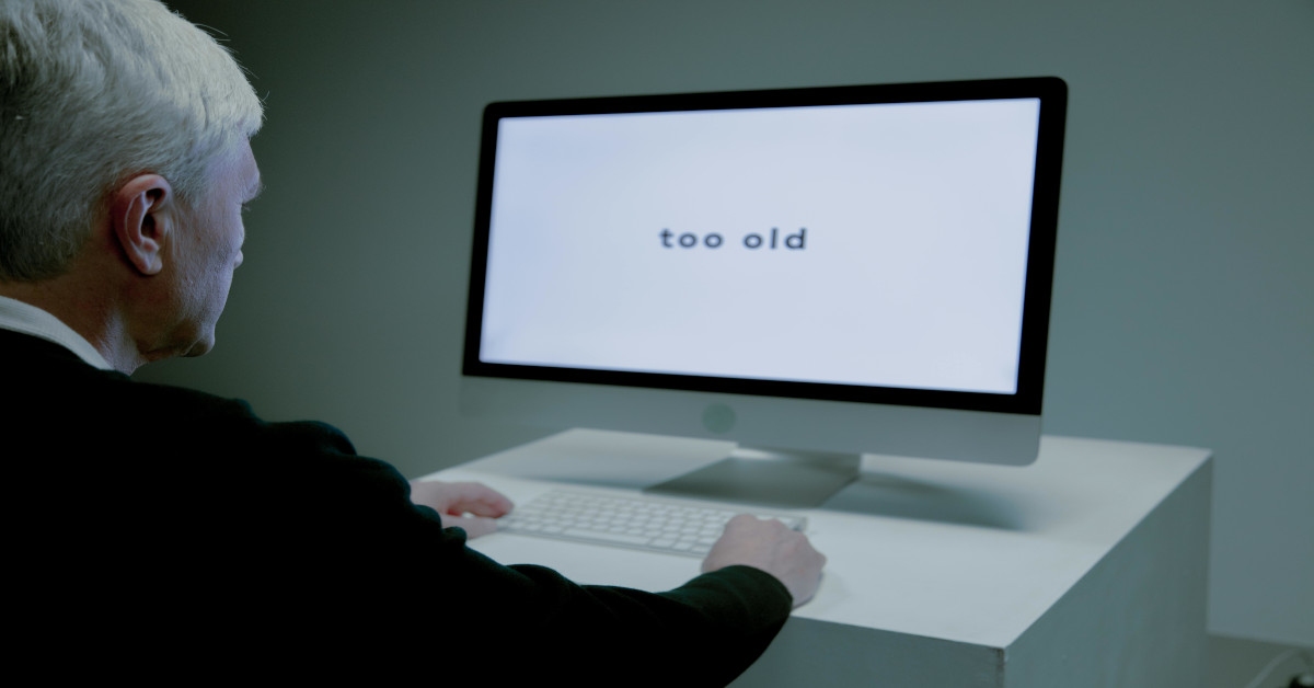 An older man is sitting in front of a computer screen that's saying "too old".