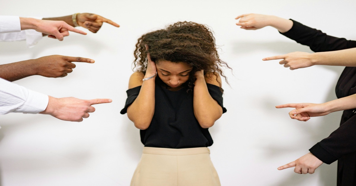 Multiple hands pointing at a woman in the middle who's putting her head down and holding her head, trying to hide.