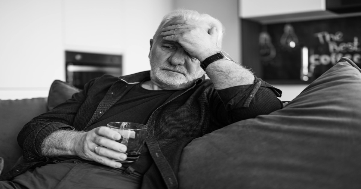 A black and white image of a sad man drinking alcohol while holding his head and sitting on the couch.
