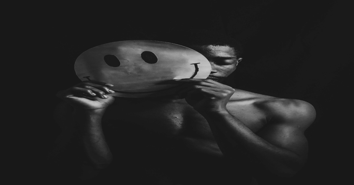 A sad man holding a smiley face in front of his face.