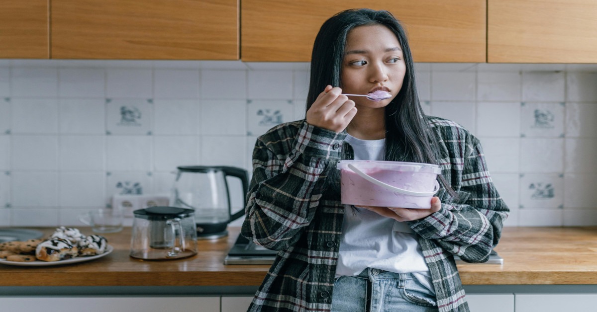 A woman is looking sad in the kitchen while eating a tub of ice cream.
