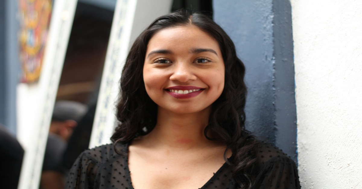 A smiling young woman is leaning against a white wall.