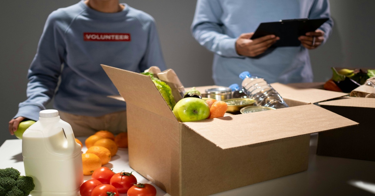 Two people volunteering by sorting food in a box while wearing a "volunteer" shirt.