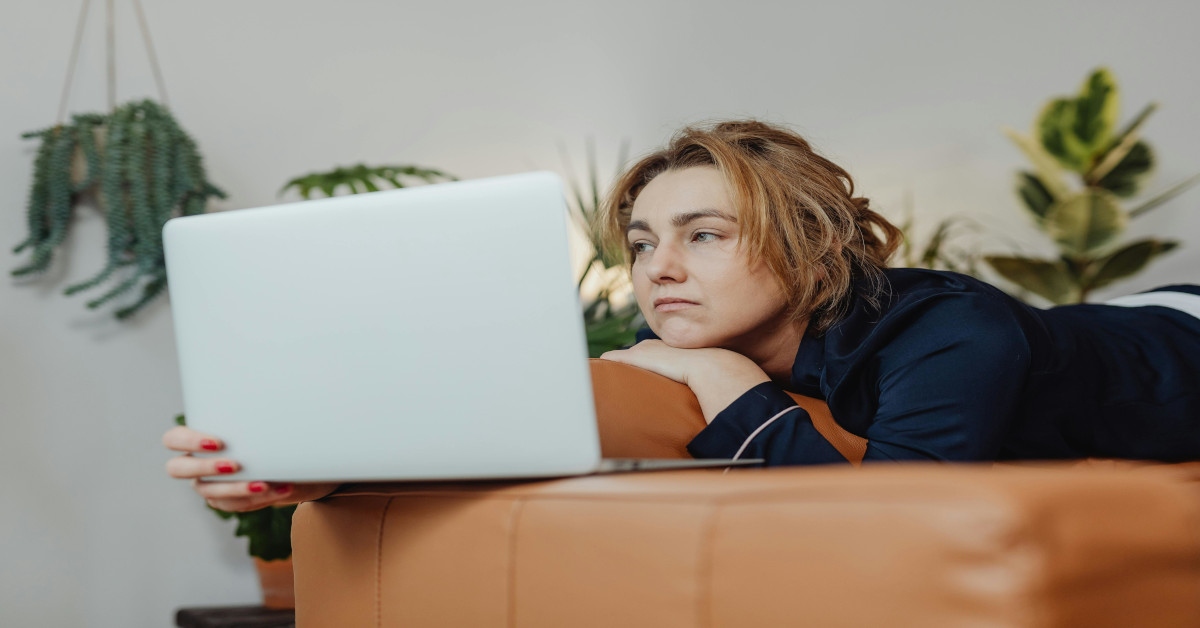 An unmotivated woman is lying on her stomach on the couch while watching her laptop and looking bored.