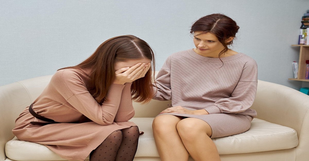 A woman is comforting another woman while both are sitting on the couch.