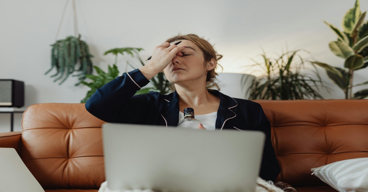 A woman drinking alcohol on the couch with a laptop on her lap while holding her head.