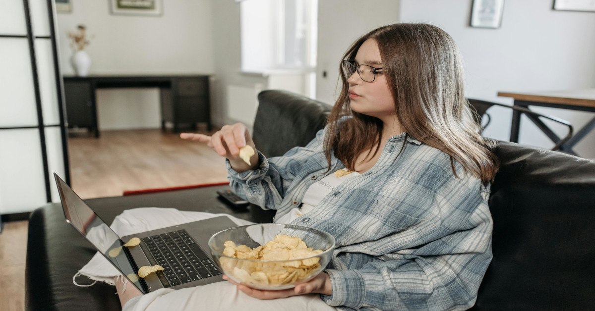 A woman eating chips while sitting on the couch and watching her laptop.