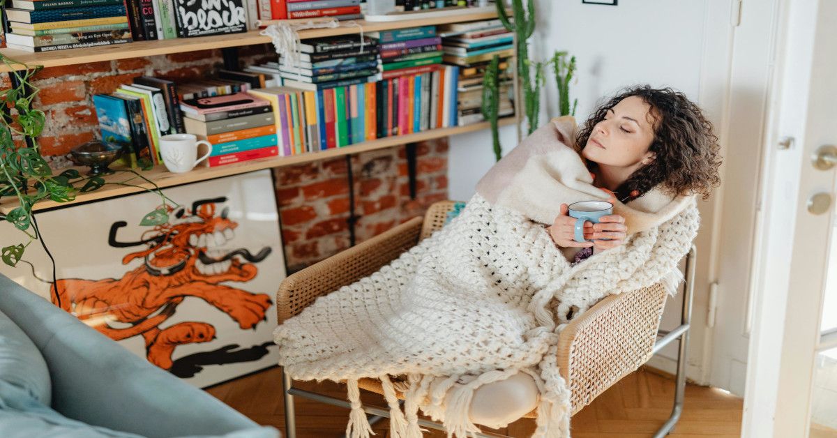 A woman feeling cold while wrapped in a blanket and holding a drink.