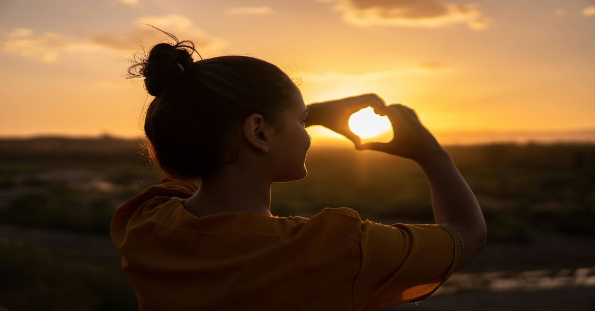 A woman making a heart sign during sunset.