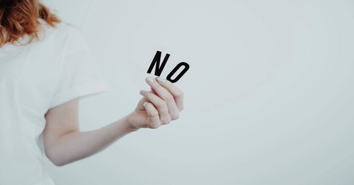A woman in a white shirt holding the word "no" in her left hand with a white background.
