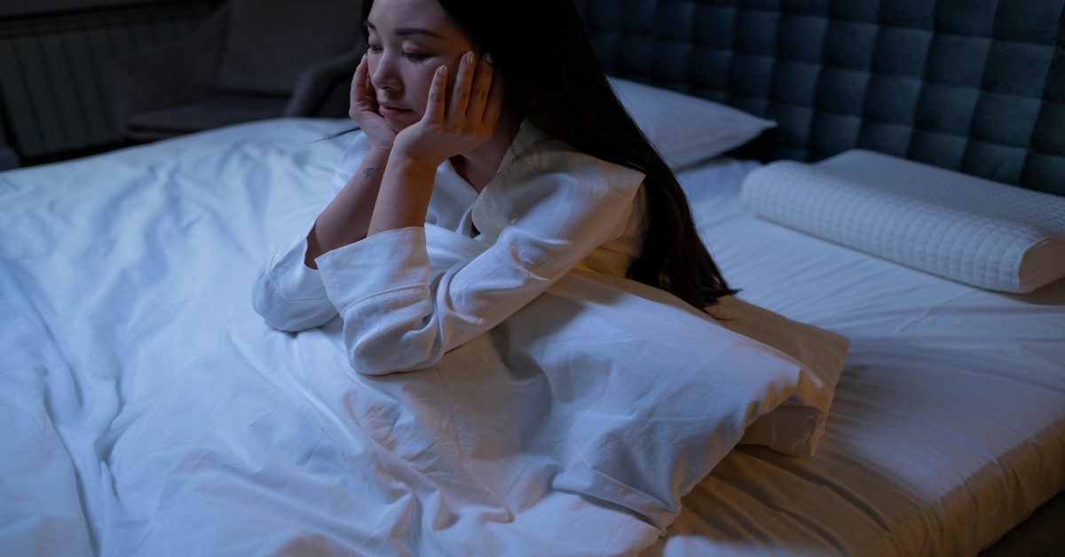 A woman is sitting upright in bed while holding her face in frustration because she can't sleep.