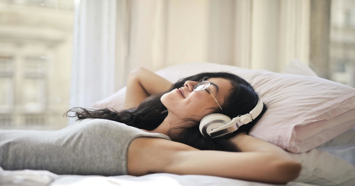 A young woman smiling while lying on her back in bed and listening to music.