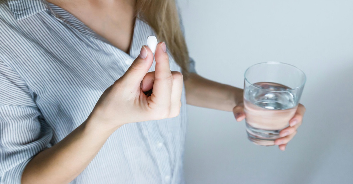A woman taking a white pill of medication with a glass of water.