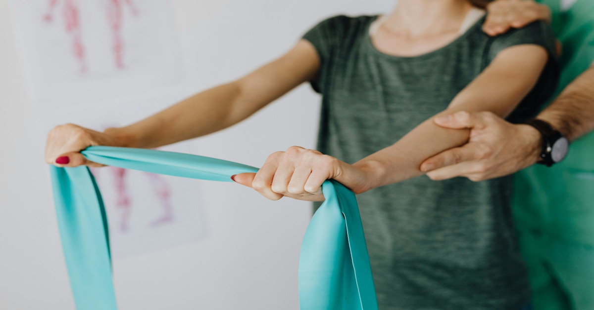 A woman stretching a resistance band under the watchful eye of a therapist.