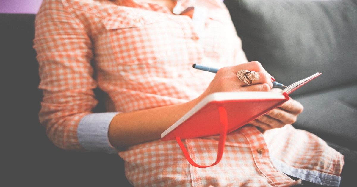 A woman writing in a red diary while sitting on the couch.