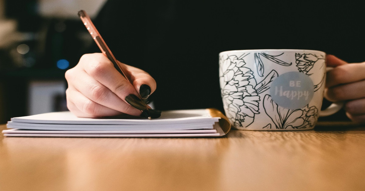 A woman is writing in a notebook while holding a coffee mug in her left hand.