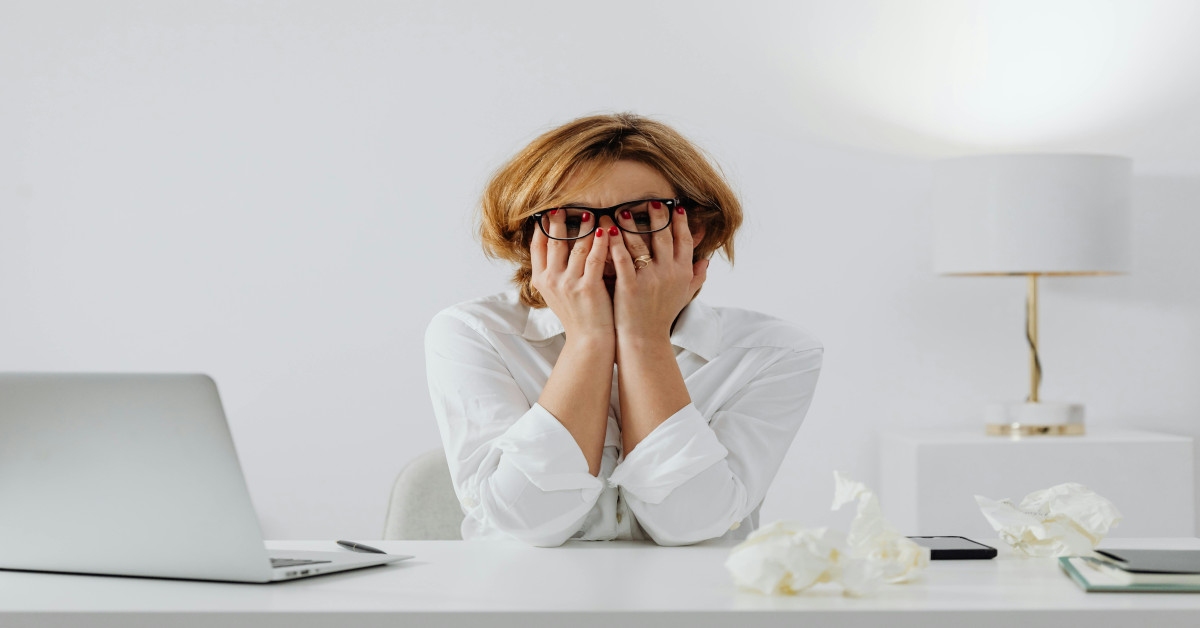 A woman holding her face in frustration behind a laptop because she's experiencing workplace stress.