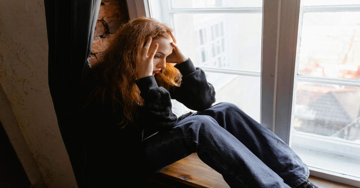 A young woman holding her head while sitting next to a window.