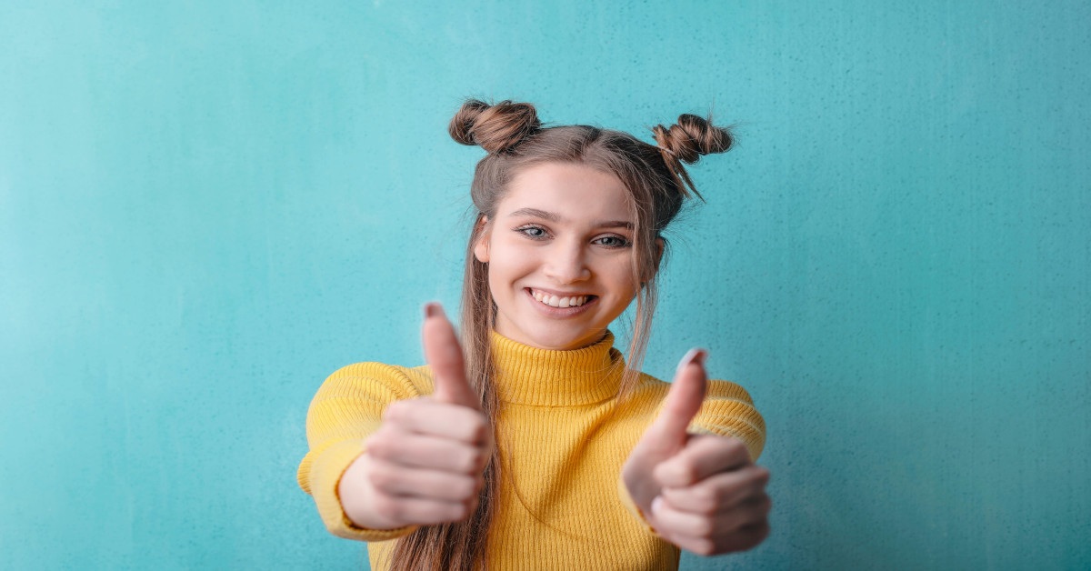 A young woman with a yellow sweater smiling while giving two thumbs up to the camera.