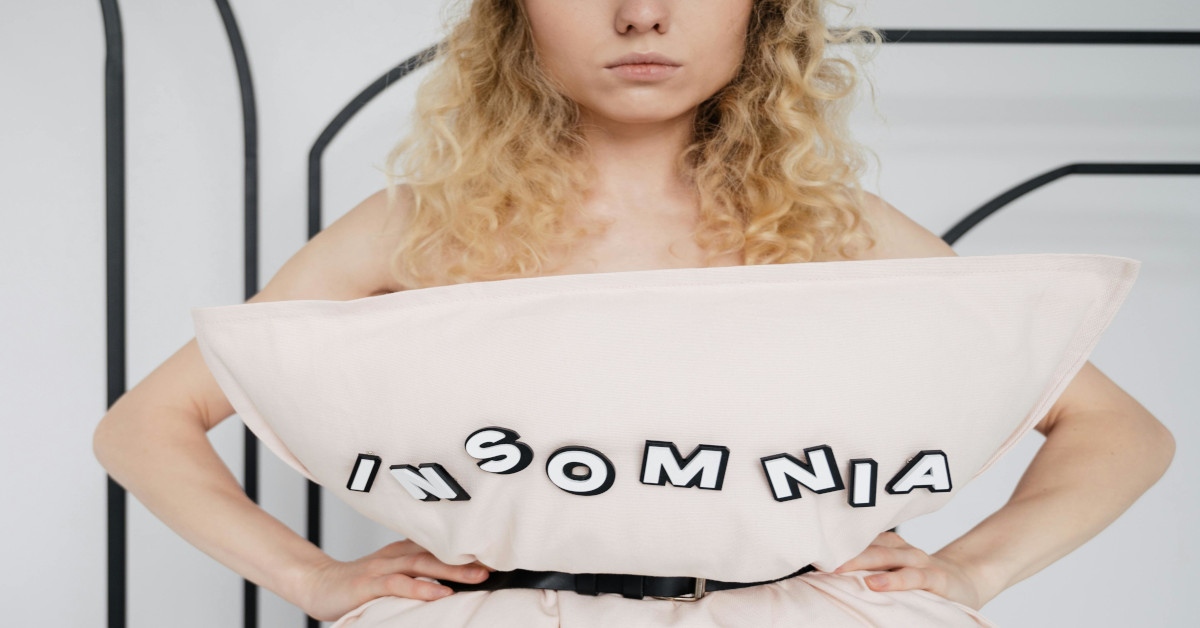 A woman holding a pillow reading "insomnia" in front of her, indicating she can't sleep.